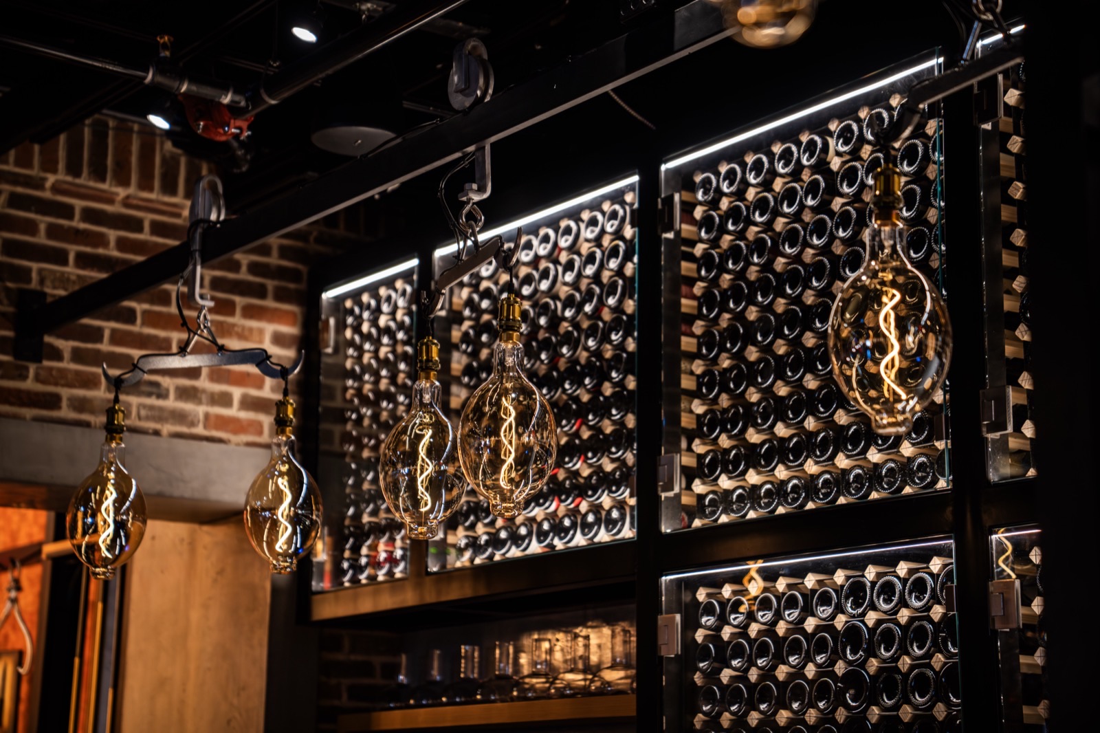 The wine wall at Little Alley Steak Alpharetta — bottles racked floor to ceiling behind glass and backlit, with oversized Edison bulbs suspended from blackened steel against exposed brick
